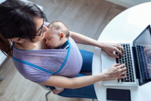 Pretty young mother with her baby in sling working with laptop at home.
